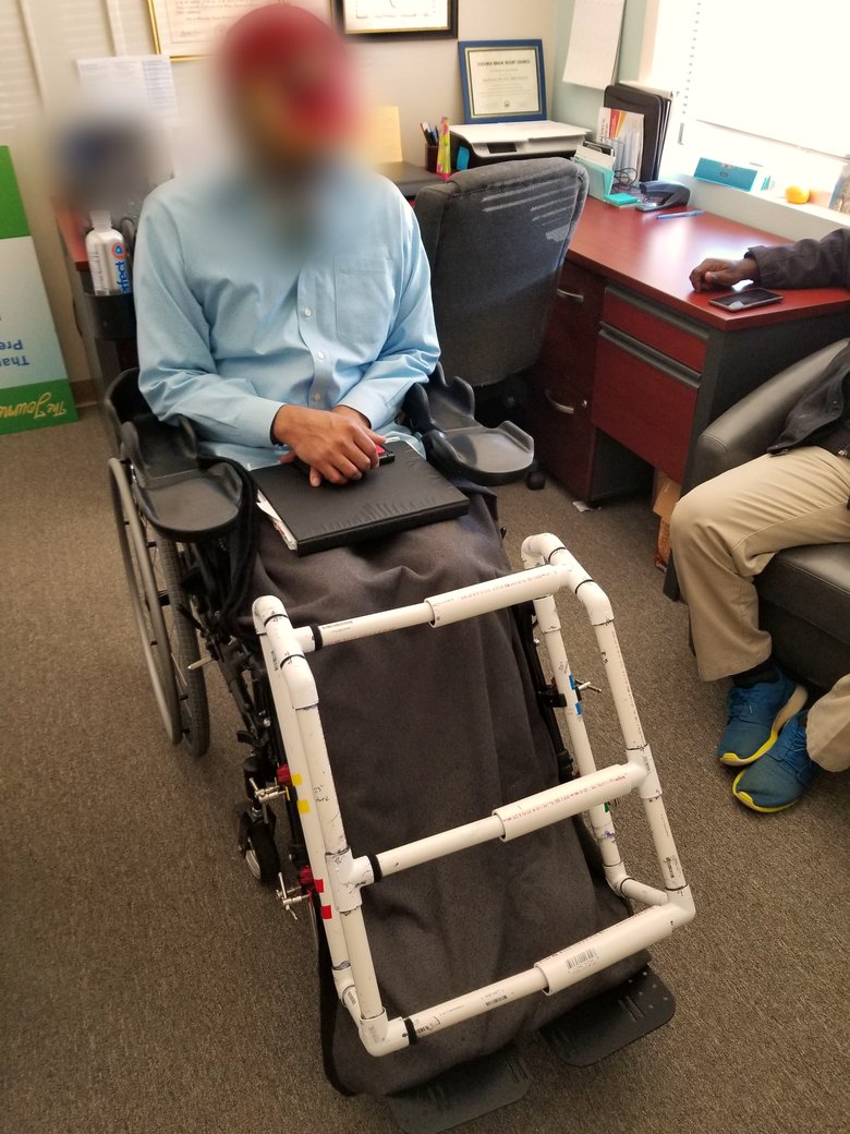 Early PVC prototype of the wheelchair leg-protector device, fitted on a client's chair during a clinical session. Subject's face anonymized. The frame uses standard PVC fittings with adjustable connectors, taped and labeled to test fit before refinement.
